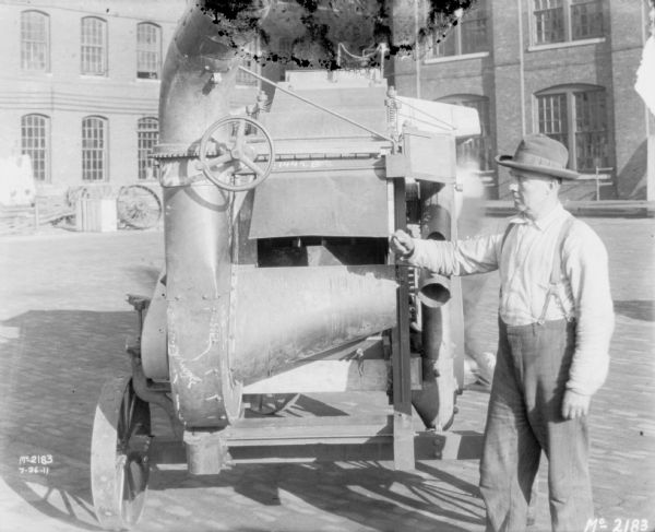 A man is standing near a Thresher outdoors in the yard at McCormick Works. A brick factory building is in the background.