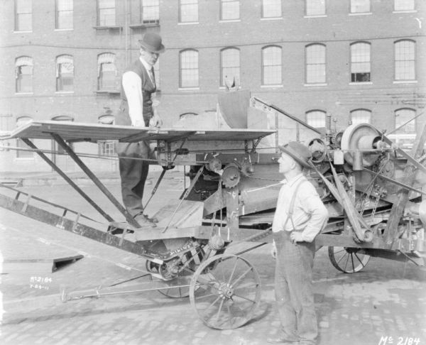 A man is standing on a thresher demonstrating the use of a thresher to another man standing in the right foreground. A brick factory building is in the background.