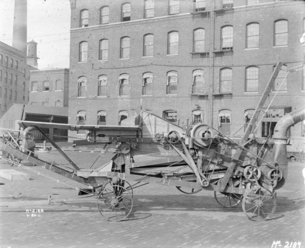 Thresher outdoors in the yard at McCormick Works. In the background are factory buildings, a smokestack, and a water tower.