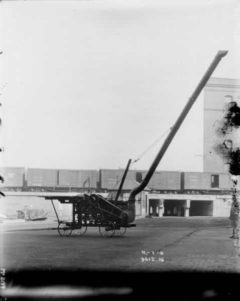 Thresher outdoors in the yard at McCormick Works. In the background are railroad cars on an elevated platform near the side of a brick factory building. A man is standing just to the right of the Thresher.