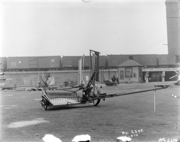 A McCormick Reaper is set up in the yard at McCormick Works. There are other pieces of agricultural machinery in the background along the side of a railroad platform, with railroad cars sitting on the tracks. There is a small wooden building near an underpass on the right.