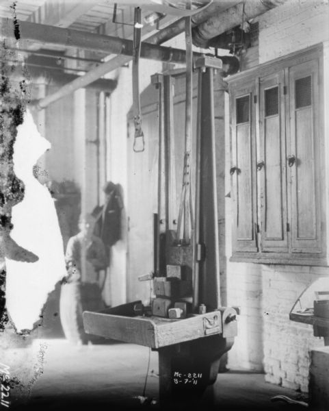 Interior view of factory, with a machine in the foreground. Cabinets are on the wall on the right. A man is standing in the background.