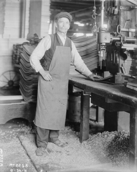 Man wearing an apron and a hat is posing standing at a drill indoors at McCormick Works.