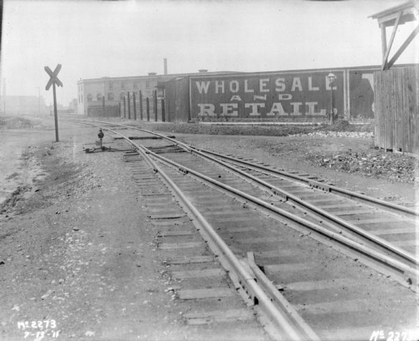 View along railroad tracks towards factory buildings. A billboard reads "Wholesale and Retail."