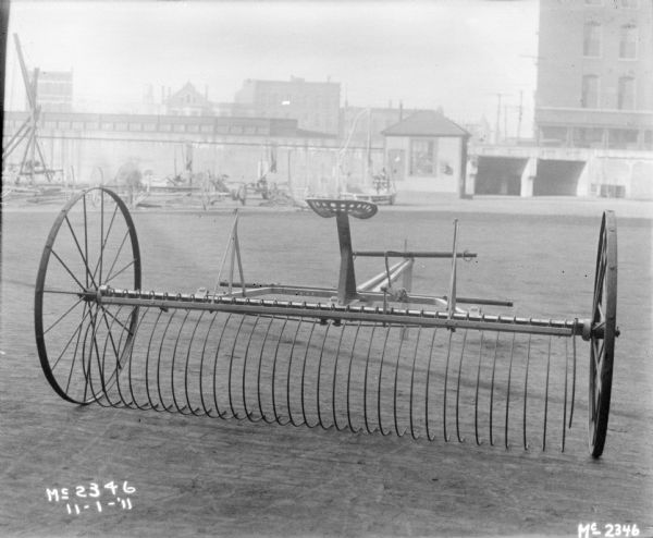 Rear view of a Dump Rake outdoors in the yard at McCormick Works. In the background is other agricultural equipment along the side of a railroad bridge. Buildings are in the background.