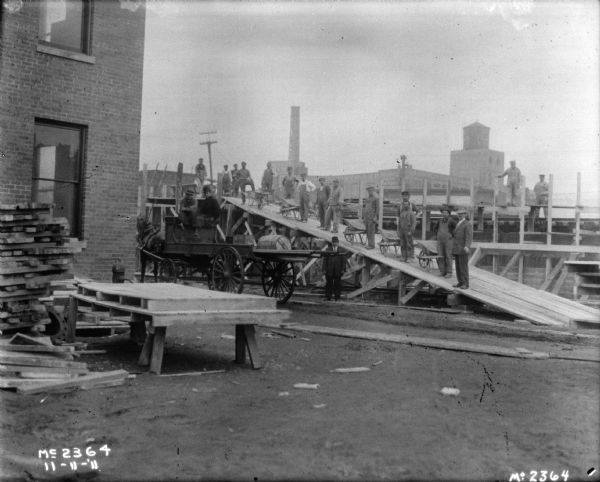 A group of employees are posing on a large wooden ramp with wheelbarrows. Two men are also posing standing wearing suits and hats. On the left is the corner of a brick factory building. Men are also posing sitting on a horse-drawn wagon on the side of the ramp on the left.