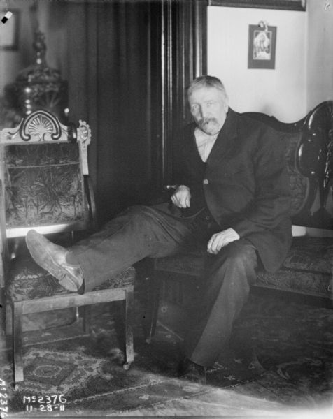 A male employee in a business suit is sitting on a couch. His right leg is resting on a chair next to him.