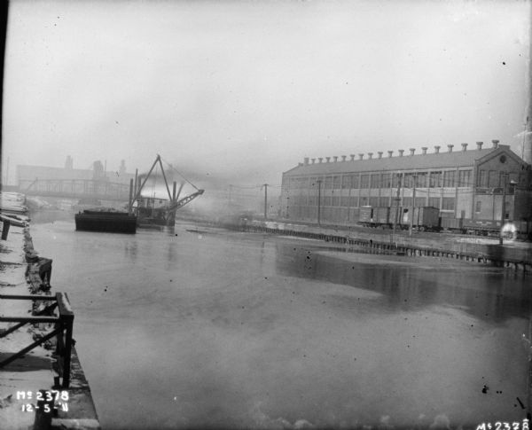 View from shipping dock down the river towards a barge, and a crane on a platform in the water. A bridge and building are in the background on the left. Across the river on the right are railroad cars and industrial buildings.