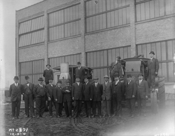 A group of men in business suits posing with two tractors. A factory building is in the background.