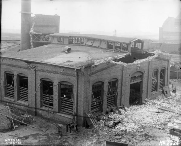 Elevated view of a collapsed roof at a factory. Bricks are scattered on the ground near a large doorway to the building.