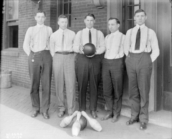 Group portrait of a men's bowling team standing against the exterior brick wall of a factory building. The man on the center is holing a bowling ball, and three pins and a bowling ball are on the ground in front of the group.