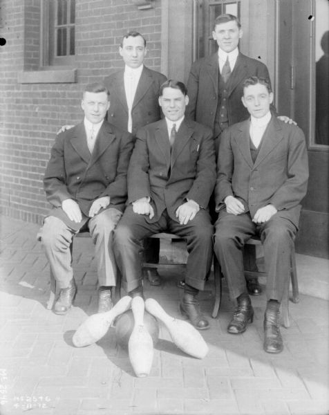 Group portrait of a men's bowling team standing against the exterior brick wall of a factory building. Three pins and a bowling ball are on the ground in front of the group.