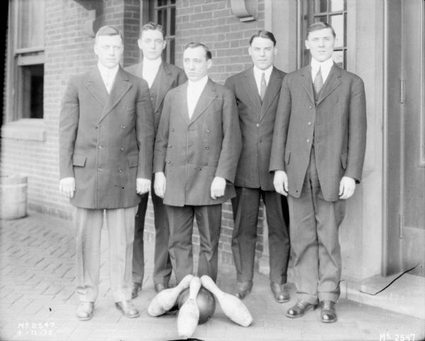 Group portrait of a men's bowling team standing against the exterior brick wall of a factory building. Three pins and a bowling ball are on the ground in front of the group.