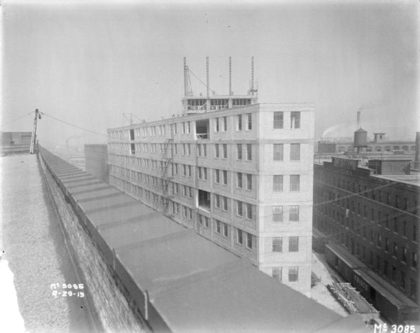 Elevated view from flat rooftop towards a wall of a factory building under construction at McCormick Works. Below on the right are railroad cars on tracks between factory buildings. A water tower is on a roof, and a sign on a roof behind the water tower reads, in reverse: "McCormick Reaper Works."