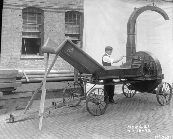 A man wearing a hat, vest and necktie is standing outdoors in the yard at McCormick Works, holding onto the side of an ensilage cutter. A brick factory building is in the background. Another man is standing behind the machinery and is holding up the side of a white cloth backdrop.