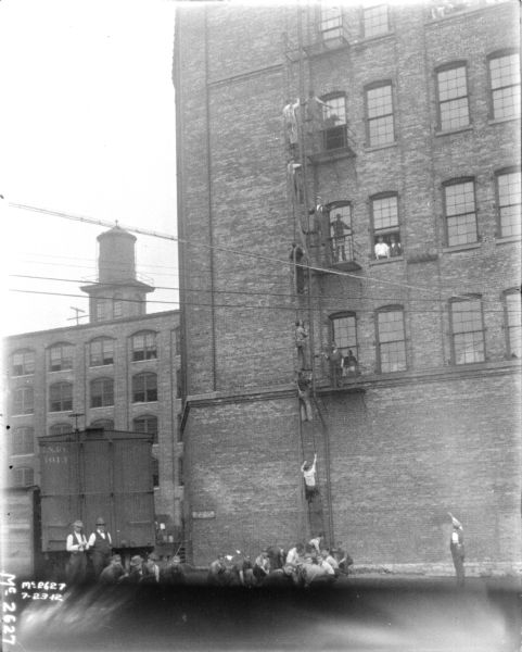 A group of employees is outdoors in front of a plant building. Other men are using the fire escape on the side of the building. A railroad car is in the background on the left.