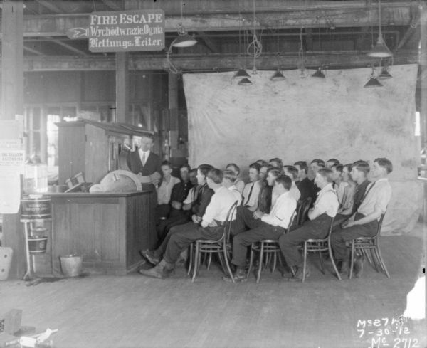 Group of men, sitting in chairs, being instructed in parts assembly. There is a backdrop hanging in the background. A sign hanging from the ceiling reads "Fire Escape" in English and two other languages.
