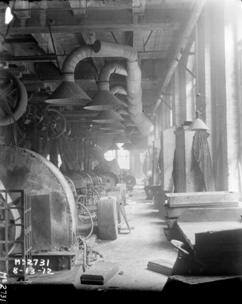 View of manufacturing area indoors. Vent hoods on the ceiling are above belt-driven machinery. Along the wall on the right are windows, and between the windows are wood cabinets with clothes hanging on the sides. Men are sitting on benches between the cabinets.