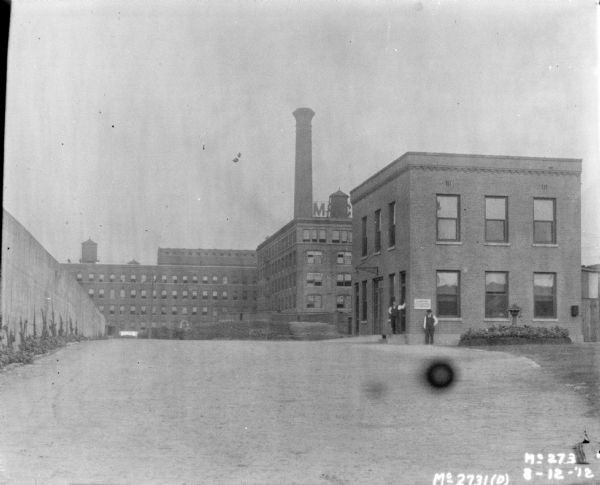 View across yard towards McCormick Works buildings. There is a wall along the left. Two men wearing hats, vests and neckties are standing near a smaller building on the right. A sign on the side of the building reads: "Smoking Prohibited."