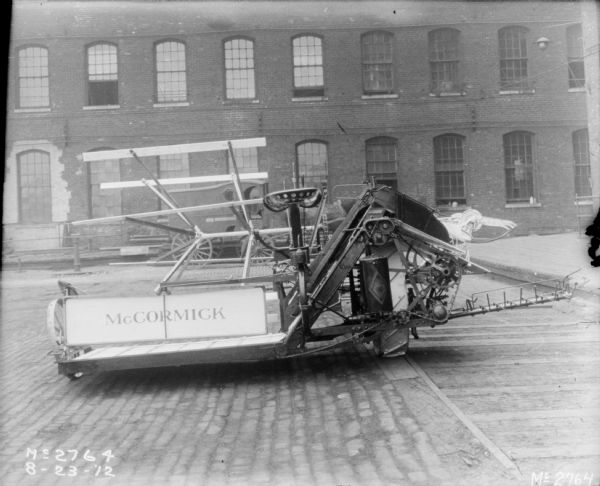 View of rear of a McCormick Binder outdoors in the yard of McCormick Works. A horse-drawn wagon in parked near a brick factory building is in the background.