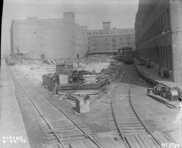 Elevated view of the yard at McCormick Works. Two sets of railroad tracks are in the foreground. Men are working among a lot of debris in the yard, and lumber and other items are stacked on a platform between the two sets of railroad tracks.