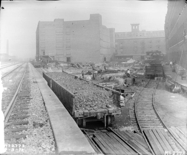 View from elevated railroad platform towards the yard with brick buildings in the background. An open railroad car on a lower set of tracks is in the foreground and is full of material. Men are working among a lot of debris in the yard, and lumber and other items are stacked on platforms.