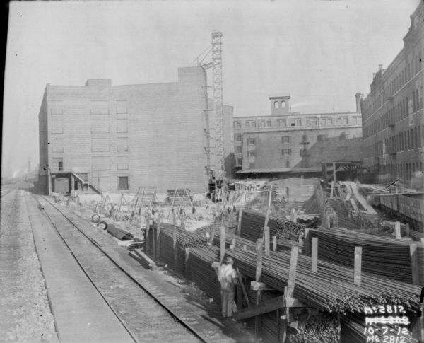 Elevated view of yard showing railroad tracks and plant construction. Men are working in the foreground near stacks of metal rods alongside the railroad tracks. There is scaffolding alongside one of the brick buildings in the background, and building materials are stacked in the yard.