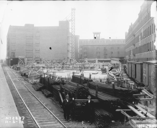 Elevated view of yard showing railroad tracks and plant construction. Two men wearing suits are standing in the foreground near stacks of metal rods alongside the railroad tracks. There are railroad cars along a factory building on the right. Scaffolding is alongside one of the brick buildings in the background, and building materials are stacked in the yard.