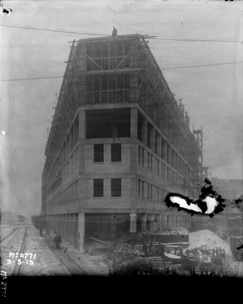 Elevated view of yard, showing construction on buildings. One man is standing on the roof of the building under construction in the center. There are men along the railroad tracks on the left.