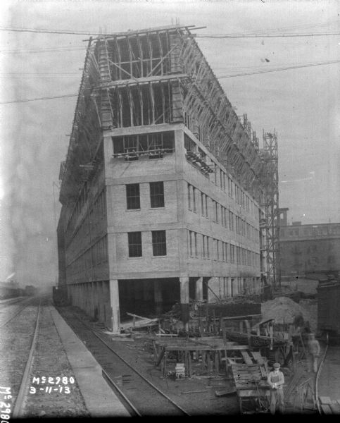 Slightly elevated view showing yard with railroad tracks and construction on buildings. Two men are standing in the right foreground. Scaffolding is alongside one of the brick buildings.