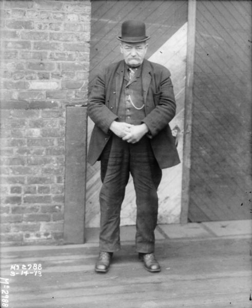 A man wearing a hat and a suit is standing outdoors near a door to a brick building.