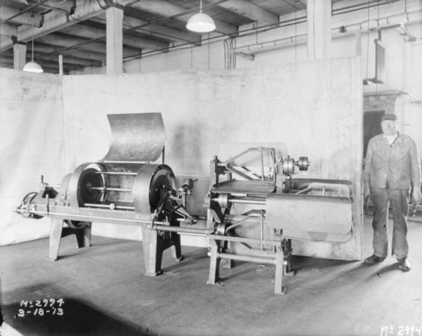 A man is standing near a manufacturing machine inside a factory building. There is a cloth backdrop set up behind the machine.