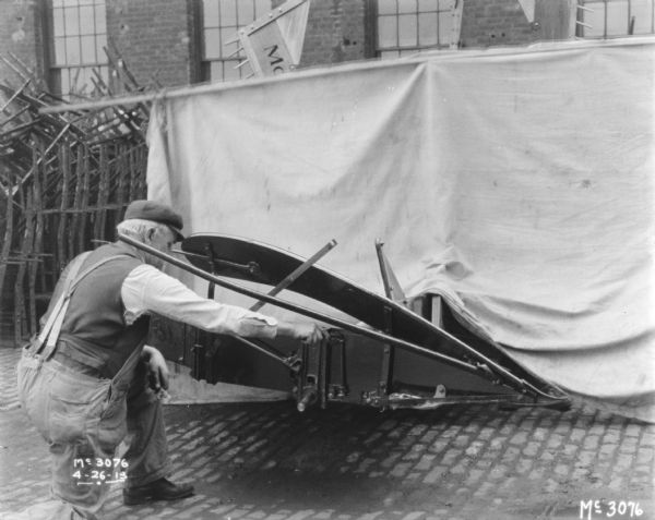 A man is adjusting a Binder outdoors in the yard. Parts are stacked behind the machine, and a brick factory building is in the background.