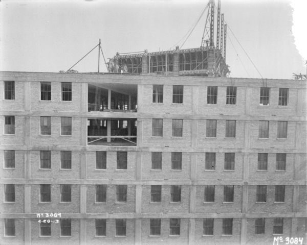 Elevated view looking towards a section of an open wall on the upper two floors of a tall building under construction. Men are standing on the flat roof above.