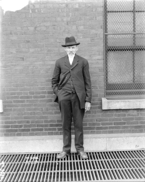 Full-length portrait of a man standing on a metal grate in front of a brick wall. A window on the right has a metal grate protecting it.