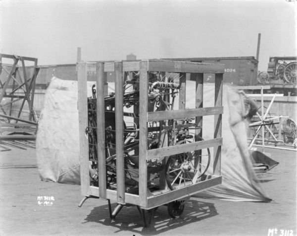 Machine crated for shipping sitting outdoors in the yard. There is a man holding up the side of a small white backdrop behind the crate. There are railroad cars on an elevated platform in the background.
