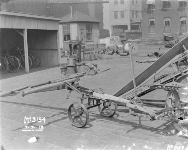 Corn binder outdoors. There are bicycles in a wooden building on the left, and the yard is cluttered with parts. Factory buildings are in the background.