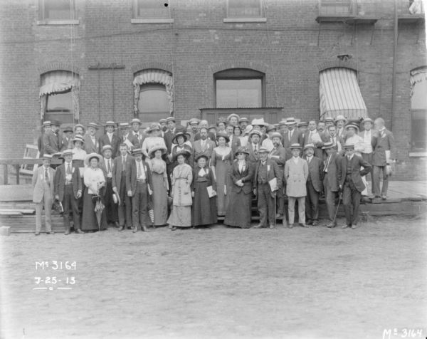 Large group of well dressed employees posing on a loading dock in front of a brick building. The women are wearing long dresses, and hats, and some are holding umbrellas. The men are wearing suits, and wearing hats, including straw boaters.
