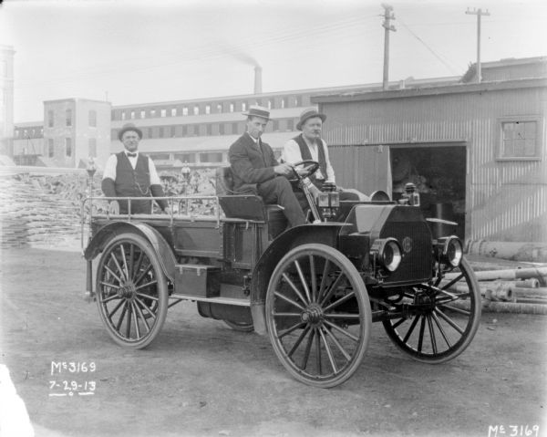 Three men are riding in a brand new fire engine. Factory buildings are in the background.
