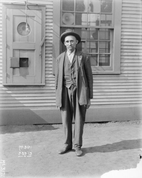 Full-length portrait of a man standing in front of a building. The ladder for a fire escape is on the wall behind him.