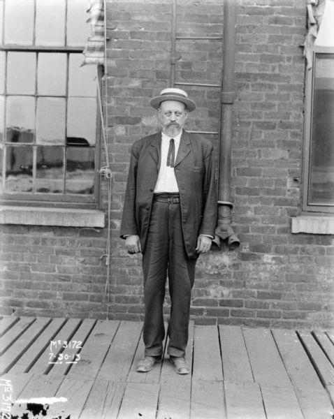 Full-length portrait of a man standing on a wood platform in front of the brick wall of a factory building. A ladder for a fire escape is on the wall behind him.