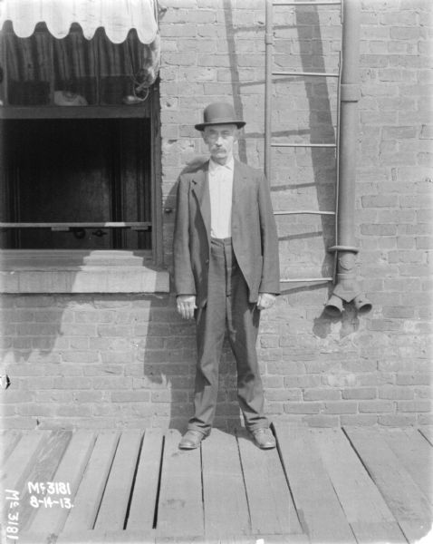 Full-length portrait of a man standing on a wooden platoform in front of the brick wall of a building. The ladder for a fire escape is on the wall behind him.