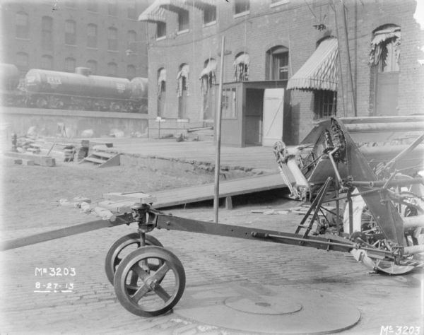 Section of a binder set up on the cobblestones outdoors in the yard. In the background is a loading dock near a brick building, and railroad tank cars on an elevated railroad platform.