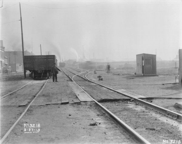 View down railroad tracks towards a man standing in front of a railroad car. There are commercial buildings along the street on the left. A train is on another set of tracks in the distance, near plant buildings.