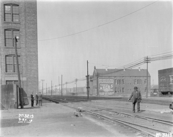 View towards multiple sets of railroad tracks running between buildings. Three men are standing next to a large brick building on the left, and another man is standing behind the railroad tracks looking at the camera and pointing down towards the tracks in front of him. Across the tracks is a large sign on a brick building that reads: "The Atlas Clothing. North-South-East-West, The Best Dressed Men Everywhere Are Wearing Our Guaranteed Clothing 1800-1808 Blue Island Ave. Corner 18th Street."