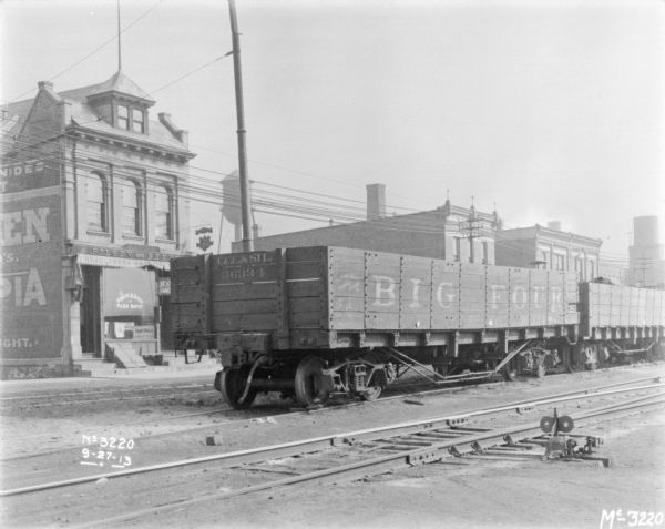 View across railroad tracks towards commercial buildings along a street. There is a wooden sided railroad car on the tracks in the foreground. A sign on the front of the building on the left reads: "Robert R. Zitek, Pilsen Buffet." There is a water tower in the backgrounc.