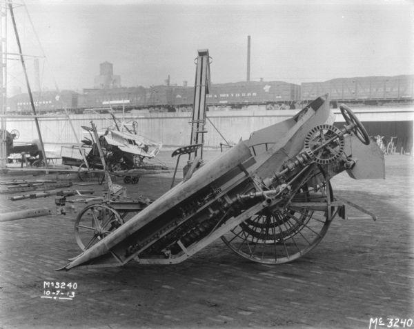 Left side view of a Corn Binder outdoors in the yard. Railroad cars on an elevated platform are in the background.