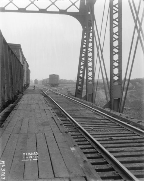 View down platform, with railroad cars on tracks on the left, and another set of railroad tracks on the right. There is a metal structure over the tracks, and a railroad yard is in the distance.