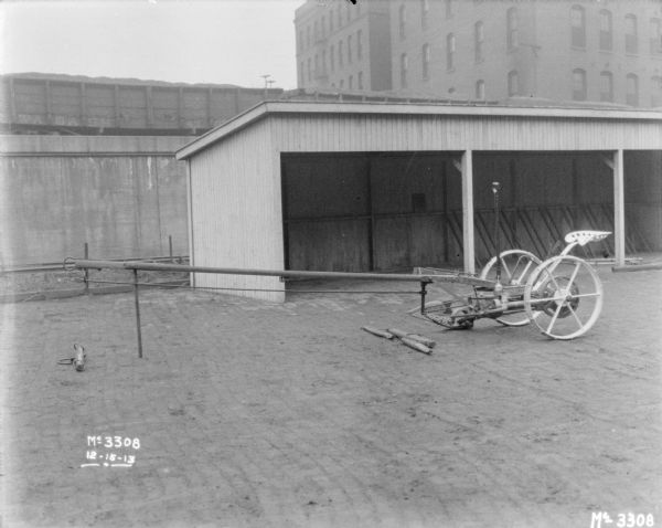 Mower outdoors on the cobblestones in the yard. Behind the mower is a three-sided shed that is empty, and railroad cars on a raised platform. Buildings are in the far background.