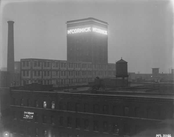 View from roof looking over factory buildings at McCormick Works. An addition on the top of one building has an electric sign near the roof that reads: "McCormick." There is a smokestack in the background on the left.